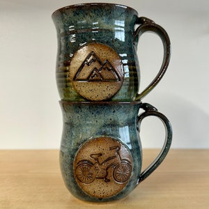 May include: Two stacked ceramic mugs with a blue-green glaze. The top mug features a mountain design, while the bottom mug has a bicycle design. Both mugs have brown handles and circular emblems.