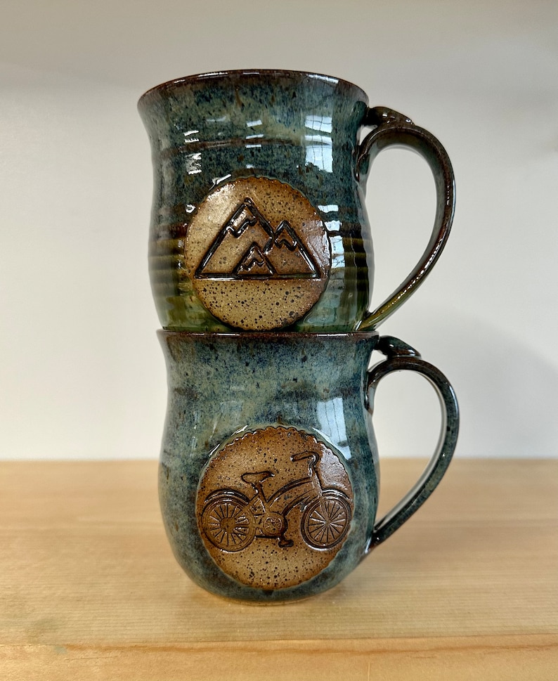 May include: Two stacked ceramic mugs with a blue-green glaze. The top mug features a mountain design, while the bottom mug has a bicycle design. Both mugs have brown handles and circular emblems.