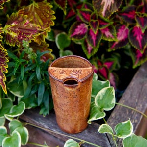 May include: A cylindrical ceramic tumbler with a speckled brown glaze and a built-in semi-circular shelf at the top. The shelf features embossed maple leaf patterns and a small opening for a tea bag string or straw, creating a functional design.