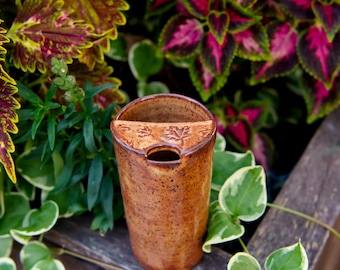 May include: A cylindrical ceramic tumbler with a speckled brown glaze and a built-in semi-circular shelf at the top. The shelf features embossed maple leaf patterns and a small opening for a tea bag string or straw, creating a functional design.