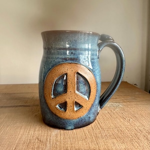 May include: A white ceramic mug with a black handle and the text "But First, Coffee" in a bold, black font. The mug is set against a blurred background, suggesting a cozy setting.