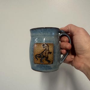 May include: Blue ceramic mug with a brown dinosaur design. The mug has a rounded shape with a handle and a textured surface. The dinosaur is embossed on a rectangular patch.