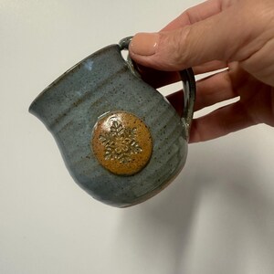 May include: A small, handmade ceramic creamer with a blue-gray glaze. It features a round, tan-colored emblem with a snowflake design. The creamer has a handle and a spout for easy pouring. The pottery has a rustic, handcrafted appearance.