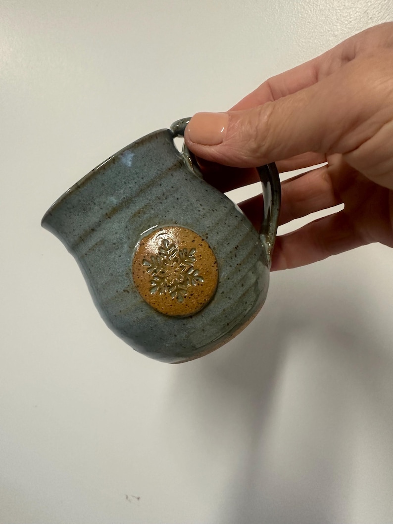 May include: A small, handmade ceramic creamer with a blue-gray glaze. It features a round, tan-colored emblem with a snowflake design. The creamer has a handle and a spout for easy pouring. The pottery has a rustic, handcrafted appearance.