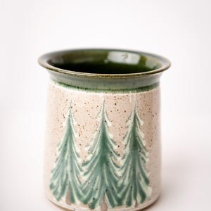 May include: A cylindrical ceramic utensil holder with a speckled beige exterior and a dark green interior and rim. Three stylized green fir trees are painted on the exterior. The holder is approximately 6 inches tall.