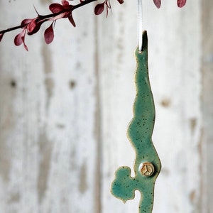 May include: A teal ceramic ornament in the shape of a person doing a yoga handstand. The ornament is speckled and has a small, round, tan-colored embellishment. It hangs from a white ribbon against a blurred, light-colored background.