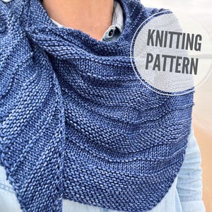 Puede incluir: Un chal de punto azul y blanco con un patrón texturizado. El chal está drapeado sobre los hombros de una persona. El texto "KNITTING PATTERN" está en un círculo blanco en el lado derecho del chal.
