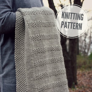 May include: A gray knitted blanket with a textured pattern. The blanket is being held by a person, and the text "Knitting Pattern" is visible in a circle in the upper right corner of the image.