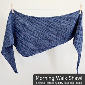 Op de afbeelding: Een blauwe gebreide sjaal met een getextureerd, geribbeld ontwerp, hangend aan een waslijn. De sjaal heeft een driehoekige vorm en is gemaakt van een zacht, geweven materiaal. De tekst "Morning Walk Shawl" wordt onderaan de afbeelding weergegeven.