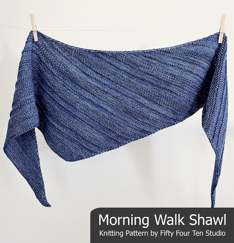 Op de afbeelding: Een blauwe gebreide sjaal met een getextureerd, geribbeld ontwerp, hangend aan een waslijn. De sjaal heeft een driehoekige vorm en is gemaakt van een zacht, geweven materiaal. De tekst "Morning Walk Shawl" wordt onderaan de afbeelding weergegeven.
