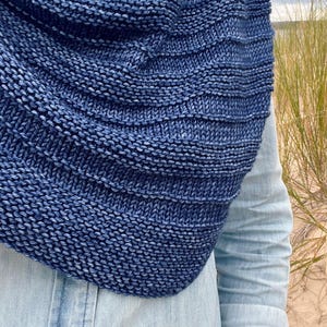 Op de afbeelding: Een close-up van een blauwe gebreide col. De col heeft een diepe indigokleur met een gestructureerd, geribbeld ontwerp. De gebreide stof lijkt zacht en warm, perfect voor koeler weer. De achtergrond omvat een lichtblauw spijkeroverhemd en duingras.