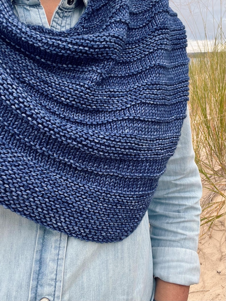 Op de afbeelding: Een close-up van een blauwe gebreide col. De col heeft een diepe indigokleur met een gestructureerd, geribbeld ontwerp. De gebreide stof lijkt zacht en warm, perfect voor koeler weer. De achtergrond omvat een lichtblauw spijkeroverhemd en duingras.