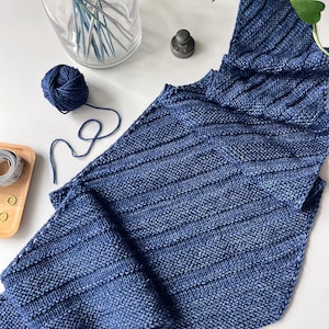 Op de afbeelding: Een blauwe gebreide sjaal met een geribbeld ontwerp. Een bol blauw garen en breinaalden zijn zichtbaar. De sjaal is opgevouwen en toont het steekpatroon en de kleurvariaties. Handgemaakt product.