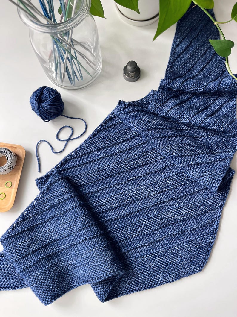 Op de afbeelding: Een blauwe gebreide sjaal met een geribbeld ontwerp. Een bol blauw garen en breinaalden zijn zichtbaar. De sjaal is opgevouwen en toont het steekpatroon en de kleurvariaties. Handgemaakt product.