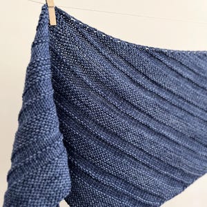 Op de afbeelding: Een marineblauwe gebreide sjaal met een geribbeld ontwerp, hangend aan een waslijn met een houten knijper. De sjaal is gemaakt van zacht, getextureerd garen en heeft een driehoekige vorm.