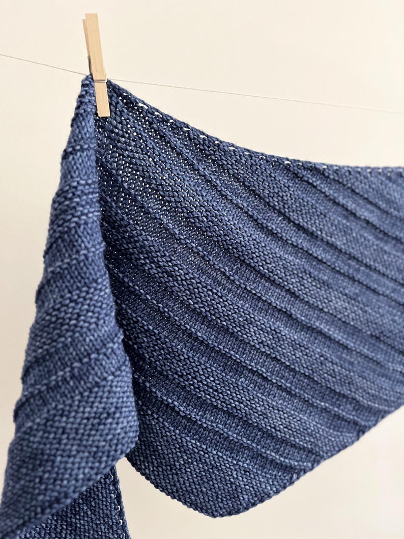 Op de afbeelding: Een marineblauwe gebreide sjaal met een geribbeld ontwerp, hangend aan een waslijn met een houten knijper. De sjaal is gemaakt van zacht, getextureerd garen en heeft een driehoekige vorm.