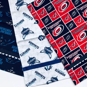 Puede incluir: Tres paneles de tela con diferentes logotipos de equipos deportivos. El primer panel es azul con texto blanco que dice "Carolina Panthers". El segundo panel es blanco con texto azul que dice "Hornets Charlotte". El tercer panel es rojo y azul con texto blanco que dice "Carolina Hurricanes".