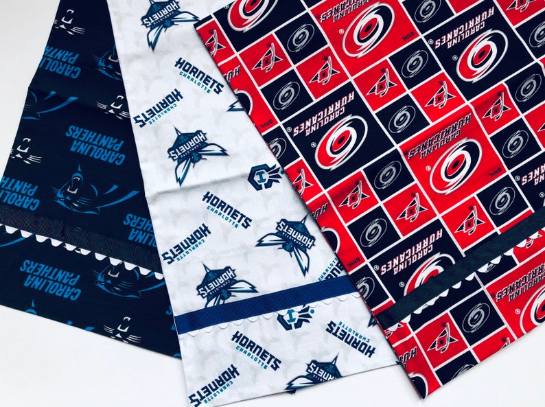 Puede incluir: Tres paneles de tela con diferentes logotipos de equipos deportivos. El primer panel es azul con texto blanco que dice "Carolina Panthers". El segundo panel es blanco con texto azul que dice "Hornets Charlotte". El tercer panel es rojo y azul con texto blanco que dice "Carolina Hurricanes".