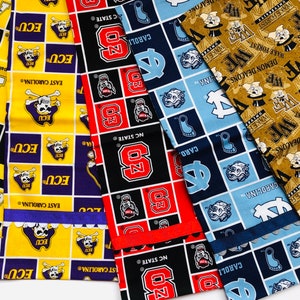 Puede incluir: Cinco cuadrados de tela con diferentes logotipos universitarios. Los cuadrados son amarillos con negro y morado, negro con rojo, azul con azul claro y dorado con negro. Los logotipos incluyen los Pirates de ECU, los Wolfpack de NC State, los Tar Heels de UNC, los Demon Deacons de Wake Forest y los Mountaineers de Appalachian State.
