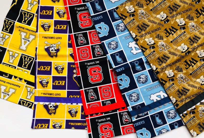 Puede incluir: Cinco cuadrados de tela con diferentes logotipos universitarios. Los cuadrados son amarillos con negro y morado, negro con rojo, azul con azul claro y dorado con negro. Los logotipos incluyen los Pirates de ECU, los Wolfpack de NC State, los Tar Heels de UNC, los Demon Deacons de Wake Forest y los Mountaineers de Appalachian State.