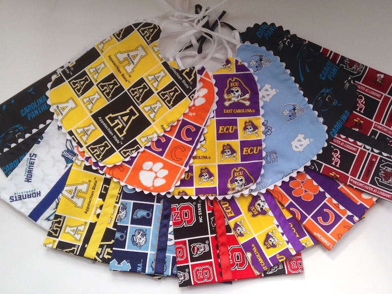 Puede incluir: Una colecci&oacute;n de 14 baberos para beb&eacute;s coloridos con varios logotipos y nombres de equipos universitarios, incluyendo Appalachian State, ECU, Carolina Panthers y NC State. Los baberos est&aacute;n hechos de tela de algod&oacute;n y tienen un borde festoneado.