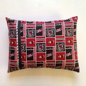 Puede incluir: Una almohada roja y negra con un patr&oacute;n repetido del logotipo de los Gamecocks de la Universidad de Carolina del Sur. El logotipo presenta un gallo con el texto "University of South Carolina" y "Gamecocks".