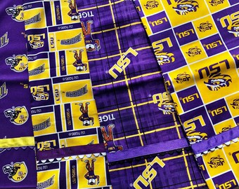 May include: Close-up of several fabric pieces featuring the LSU Tigers logo and colors. The fabrics are primarily purple and gold, with repeating patterns of the team's logo, the letters "LSU", and the word "Tigers". Some pieces have decorative edges.