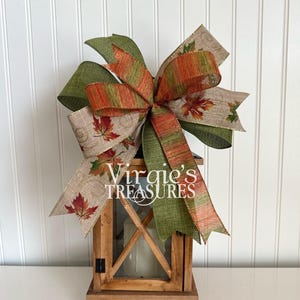 May include: A wooden lantern with a large decorative bow. The bow features green, orange, and tan ribbons with fall leaf patterns. The lantern has a rustic design with a candle inside. The text "Virgie's TREASURES" is visible on the bow.