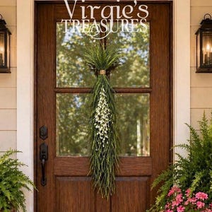 May include: A wooden door with a decorative green and white floral swag. The door has a glass window with the text "Virgie's TREASURES" in white. Black lanterns are on either side of the door, and potted plants with pink flowers are in the foreground.