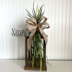 May include: A wooden lantern with a burlap bow and green leafy garland on top. The lantern is sitting on a white surface. The text "Virgie's Treasures" is visible in the image.