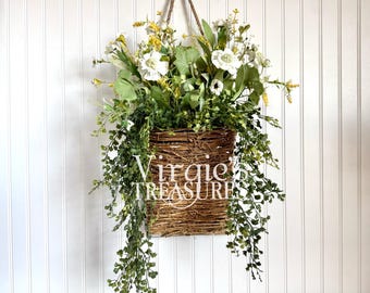 May include: A woven basket filled with white and yellow flowers and green foliage, hanging from a rope. The basket has the text "Virgie TREASURE" printed on it. The basket is hanging against a white, vertical-slat wall.