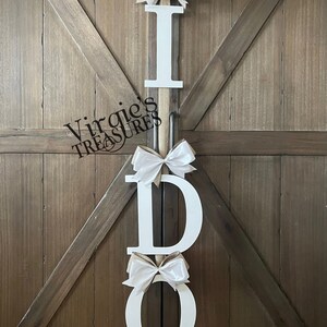May include: White wooden letters spelling "I DO" with white satin bows on each letter. The letters are hanging on a wooden door.
