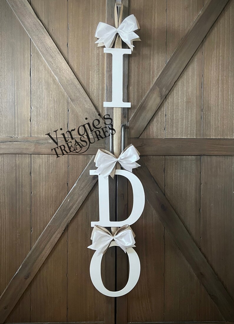 May include: White wooden letters spelling "I DO" with white satin bows on each letter. The letters are hanging on a wooden door.