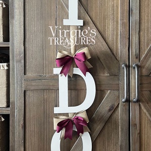 May include: White wooden letters spelling "I DO" with burlap and burgundy bows hanging on a wooden door. The letters are decorated with burlap and burgundy bows. The text "Virgie's Treasures" is visible below the top letter.