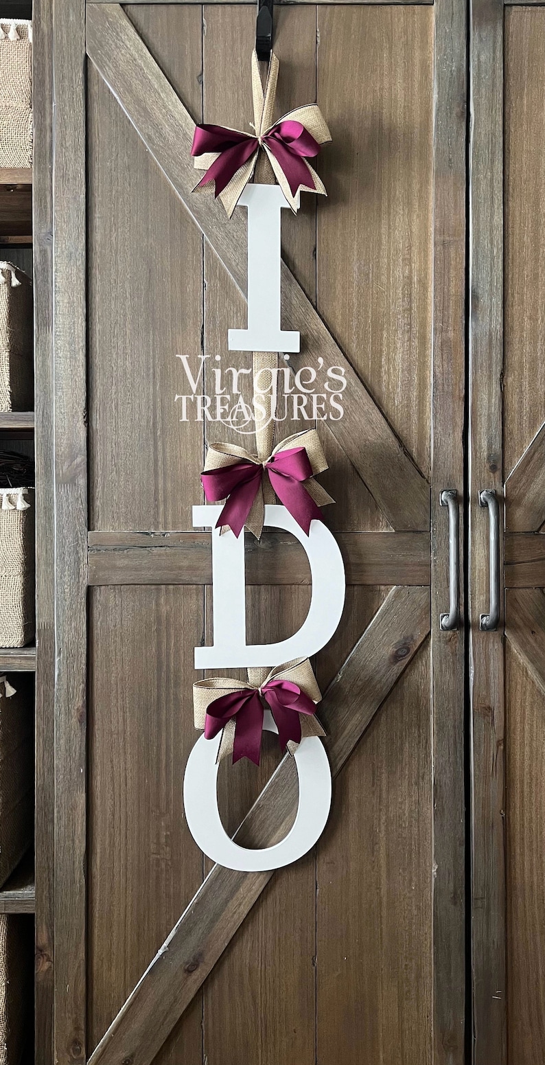 May include: White wooden letters spelling "I DO" with burlap and burgundy bows hanging on a wooden door. The letters are decorated with burlap and burgundy bows. The text "Virgie's Treasures" is visible below the top letter.