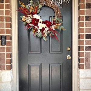May include: A burgundy and white floral wreath with greenery and natural elements hangs on a black front door. The wreath is decorated with a sign that reads "Virgie's Treasures".