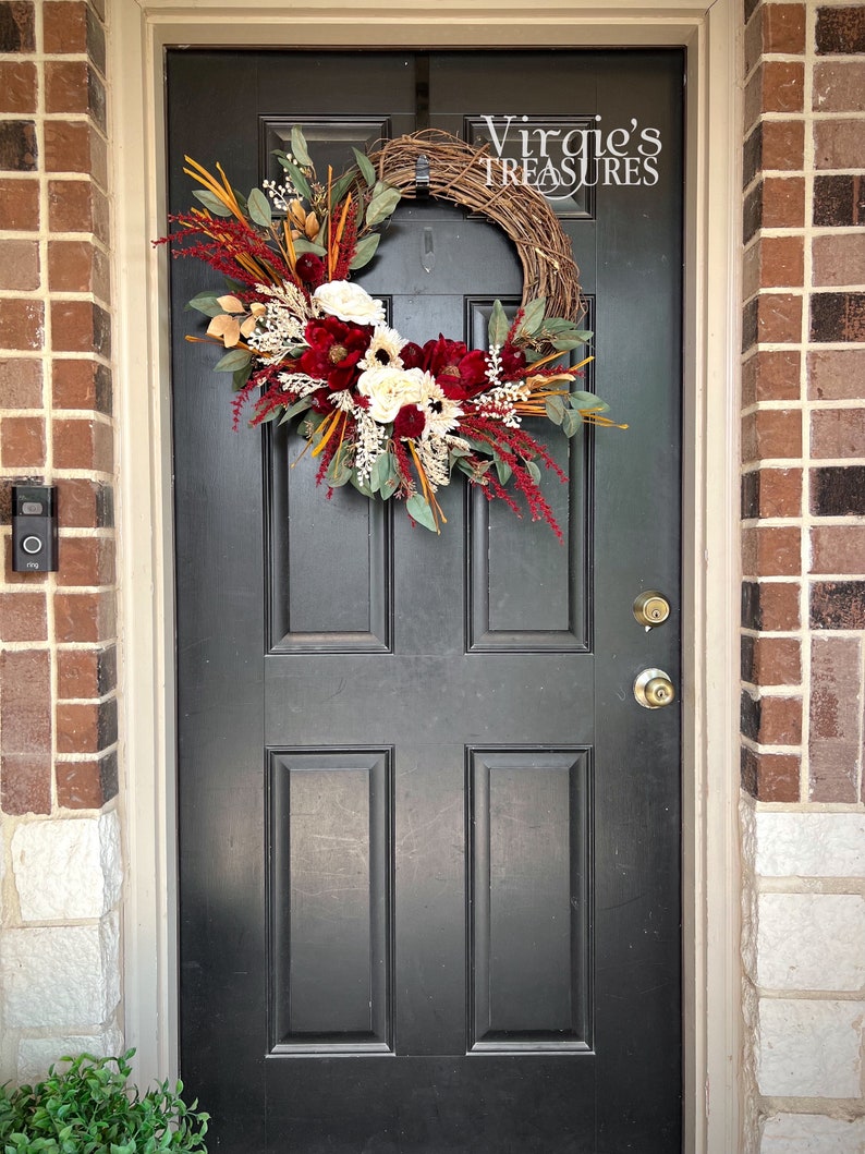 May include: A burgundy and white floral wreath with greenery and natural elements hangs on a black front door. The wreath is decorated with a sign that reads "Virgie's Treasures".