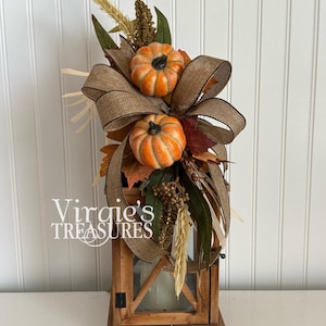 May include: A wooden lantern topped with a decorative arrangement featuring two orange pumpkins, a large brown burlap bow, dried wheat stalks, green leaves, and brown berries. The text "Virgie's TREASURES" is printed in white on the left side.