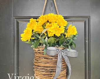 May include: A yellow flower wreath hanging on a black door. The wreath is made of a woven basket with a black and white striped ribbon.