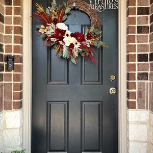 May include: A black front door with a fall-themed wreath featuring burgundy, white, and gold flowers, greenery, and berries. The wreath is hanging on a black door with a gold door handle. The text "Virgie's Treasures" is visible on the wreath.