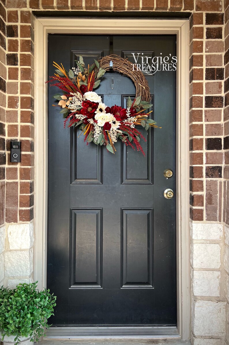 May include: A black front door with a fall-themed wreath featuring burgundy, white, and gold flowers, greenery, and berries. The wreath is hanging on a black door with a gold door handle. The text "Virgie's Treasures" is visible on the wreath.