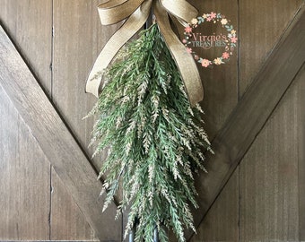 May include: A teardrop-shaped swag featuring green and white faux cedar foliage, topped with a large, multi-looped tan burlap bow with dark edges. The text Virgie's Treasures is visible in a floral wreath logo on the upper right side.