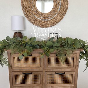 May include: A wooden dresser with four drawers and woven cane fronts. The dresser has black handles and is decorated with a eucalyptus garland. A white lamp with a brown base sits on top of the dresser. A round woven mirror hangs on the wall above the dresser. The text "Virgie's Treasures" is visible on the wall.
