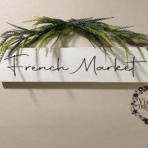 May include: White wooden sign with black cursive text that reads "French Market". A green leafy garland hangs above the sign.