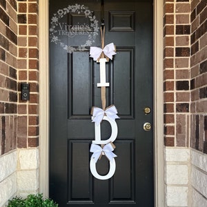 May include: A white wooden "I DO" sign with white bows hanging on a black front door. The sign is decorated with burlap ribbon.