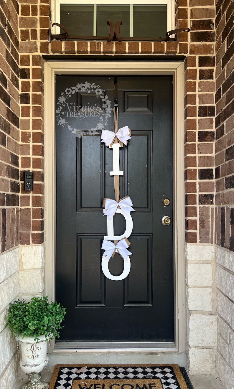 May include: A white wooden "I DO" sign with white bows hanging on a black front door. The sign is decorated with burlap ribbon.