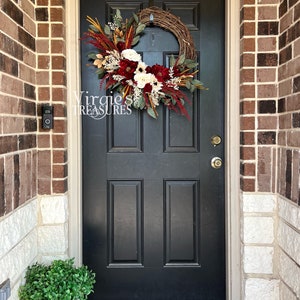 May include: A black front door with a fall-themed wreath featuring burgundy, white, and cream flowers, greenery, and dried grasses. The wreath is hanging on a brick wall with a white stone trim. A small potted plant sits on the ground in front of the door.