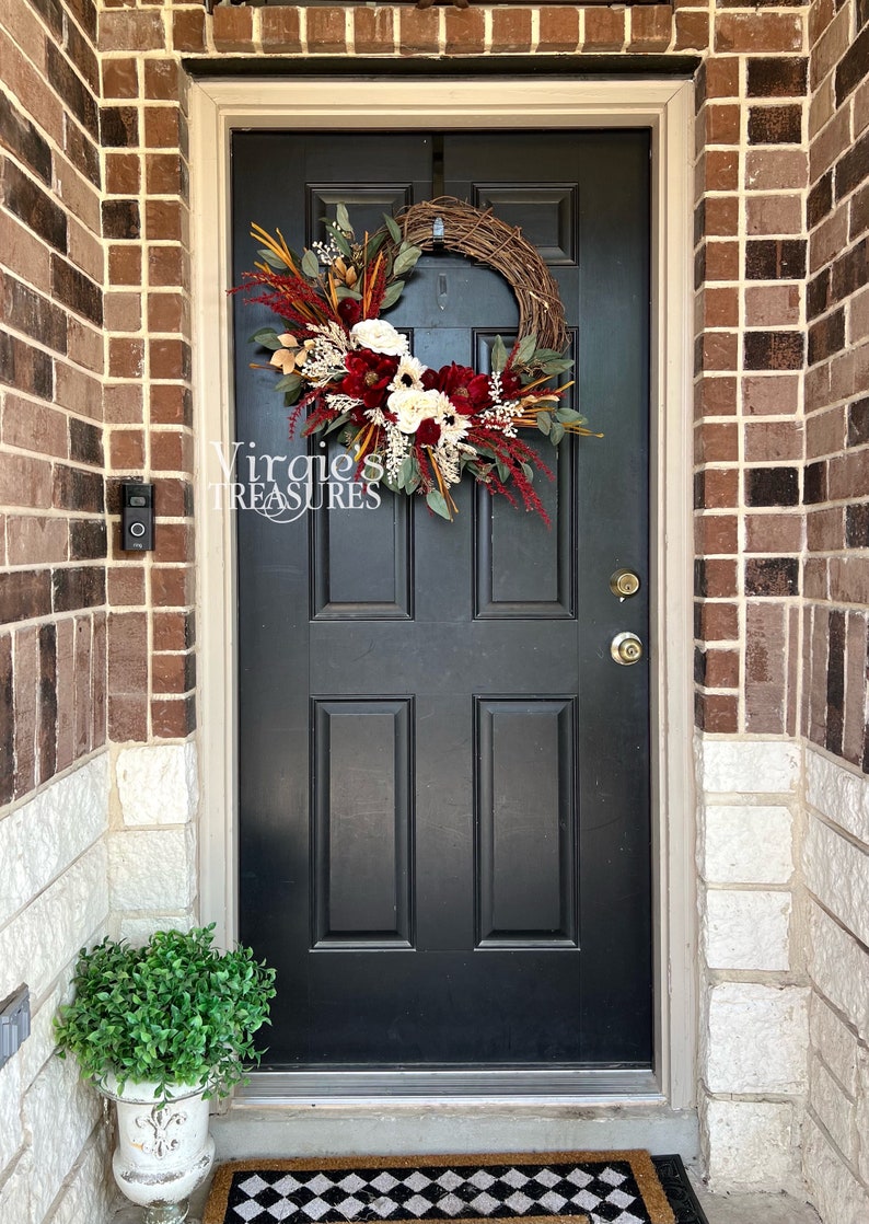 May include: A black front door with a fall-themed wreath featuring burgundy, white, and cream flowers, greenery, and dried grasses. The wreath is hanging on a brick wall with a white stone trim. A small potted plant sits on the ground in front of the door.