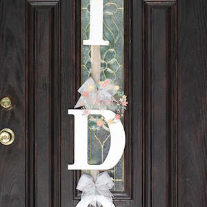 May include: White wooden letters spelling "I DO" with a white lace bow and floral accents hanging on a dark brown wooden door.