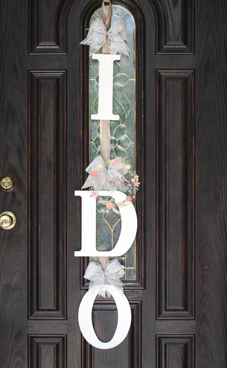 May include: White wooden letters spelling "I DO" with a white lace bow and floral accents hanging on a dark brown wooden door.
